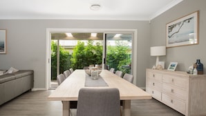 A modern dining room with a long table, grey upholstered chairs, a wooden cabinet, and a view of a lush garden through glass doors. - Callala Bay Beach House - Wowly Waters - Pet Friendly 🐳 (Callala Bay)