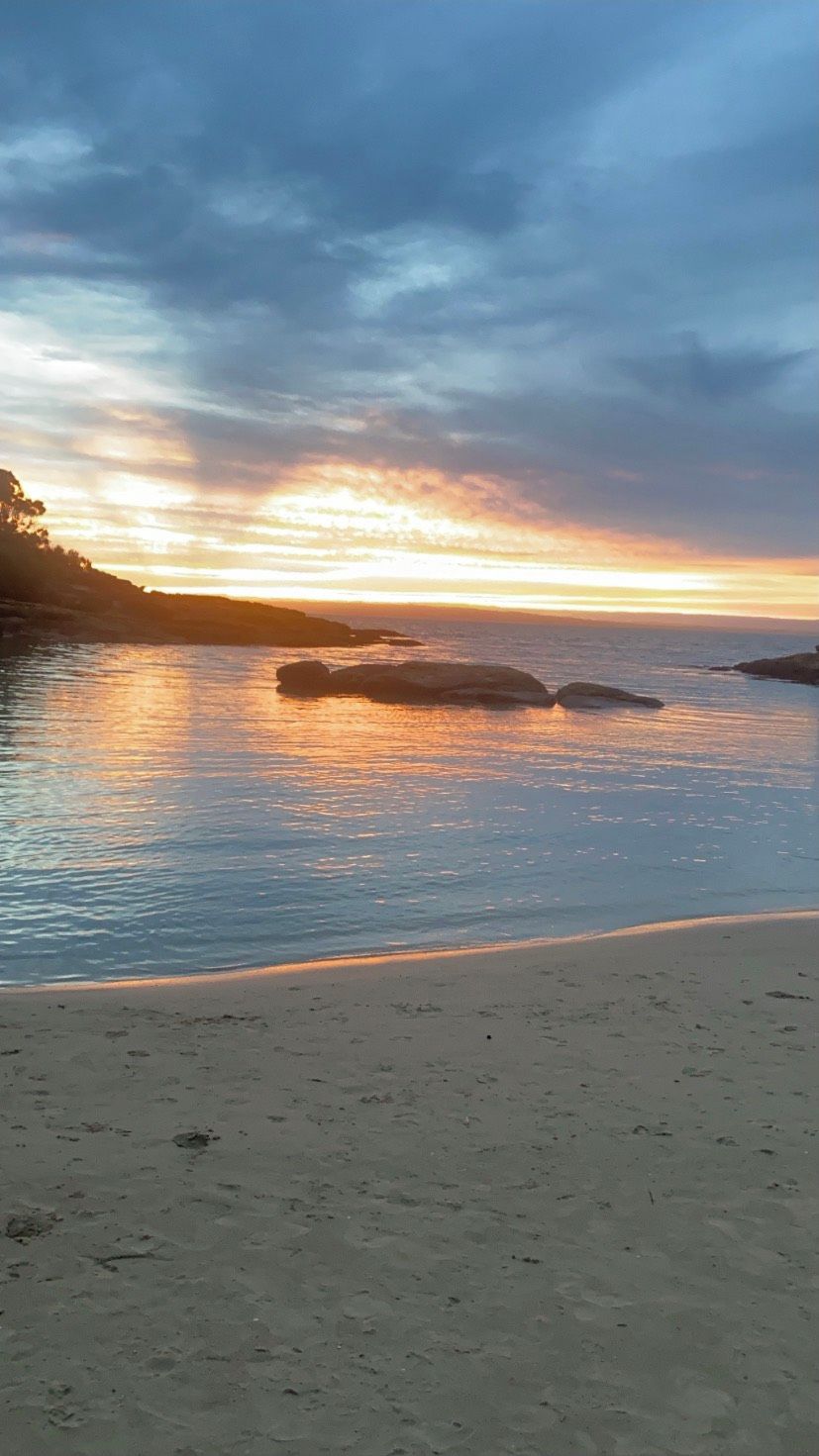 A beach with calm water, a sandy shore, and a sunset in the background.