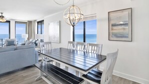A modern dining area with a dark wooden table and white chairs, large windows with a view of the ocean, and a chandelier. - Beachfront 3BR @ Phoenix X • Sleeps 10 • Gulf Views • Next to Flora-Bama & Resort Amenities (Orange Beach)