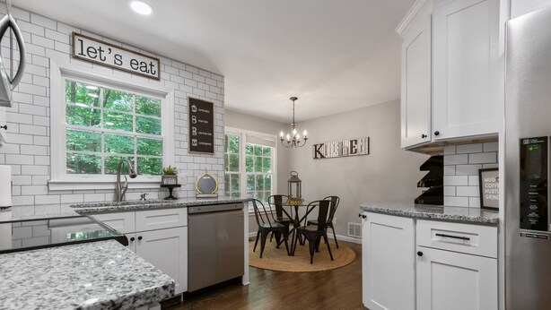 A modern kitchen with white cabinetry, granite countertops, and a central island. There are two windows providing natural light, and a sign on the wall that reads "let's eat".