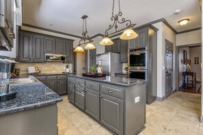 Une cuisine avec des meubles gris, un plan de travail en granit, des appareils électroménagers en acier inoxydable et des luminaires suspendus.