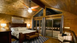 Ein gemütliches, helles Schlafzimmer mit einem großen Bett, einem Holzkopfteil, einem Deckenventilator und Blick ins Freie durch große Fenster.