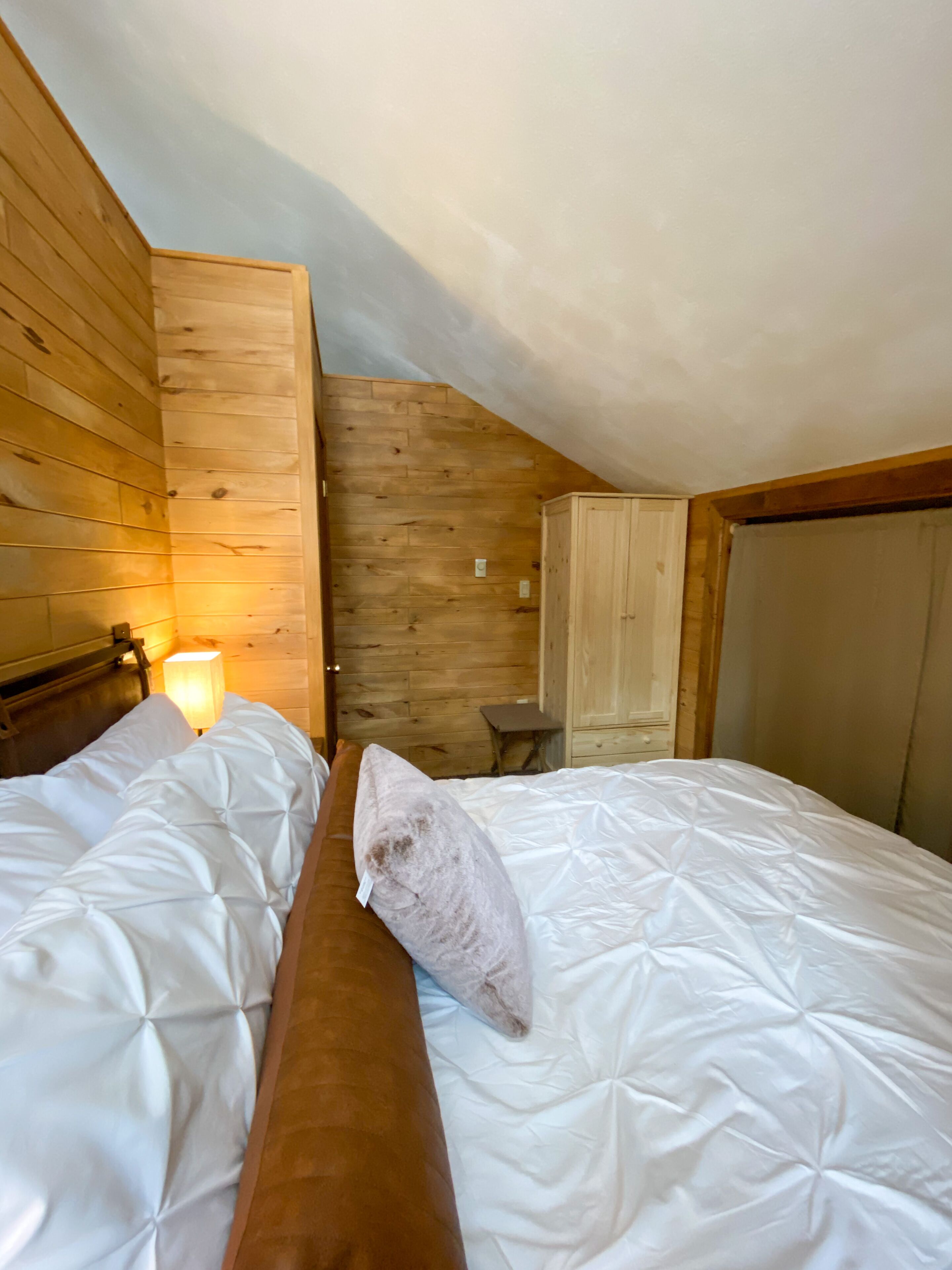 Un dormitorio con dos camas, paredes de madera, un banco de madera, una mesita de noche con lámpara y un armario.