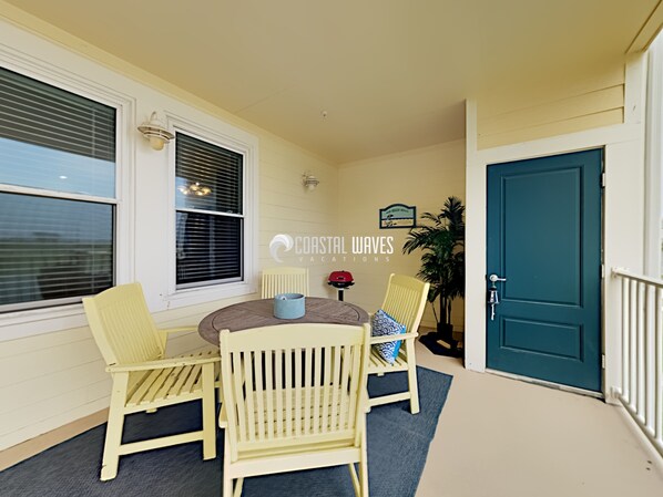 A covered patio with a round table and four chairs, a potted plant, and a door. - Pearl Life | Spacious Coastal Condo | Sleeps 10 (Galveston)