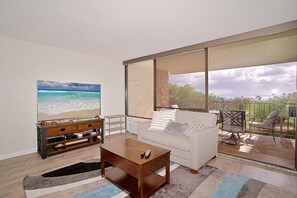 Una sala de estar con televisor grande, un sofá, una mesa de centro y un balcón con mobiliario exterior.