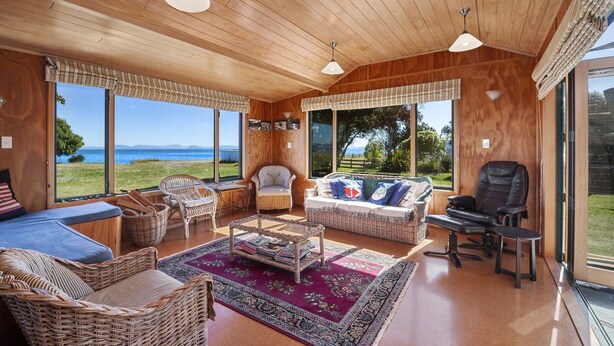A cozy room with wicker furniture, a sofa, and a chair, large windows with a view of the sea, and a wooden ceiling.