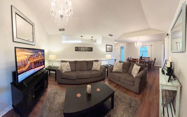 A living room with a large flat-screen TV, two sofas, a coffee table, and a dining area in the background.