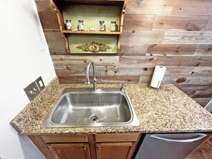 A kitchen with a stainless steel sink, wooden cabinets, a granite countertop, and a wooden wall with a shelf above the sink.