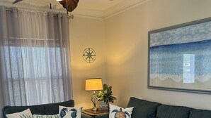 A living room with a ceiling fan, a couch with beach-themed pillows, a side table with a lamp, and a framed picture of the ocean. - Condo super close to the beach with a view of SpaceX Launch in the sky! (south padre island)