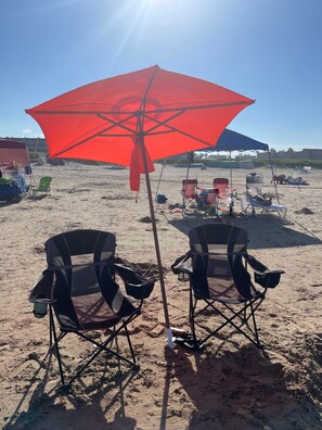 A beach scene with two black folding chairs under a red umbrella, a beach chair cart, and a tent in the background.