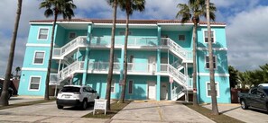 A turquoise multi-story building with balconies, parked cars, and palm trees.
