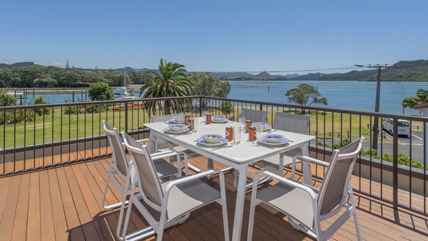 Mesa de comedor con sillas en una terraza de madera con vista a un lago con barcos, rodeado de vegetación y colinas.