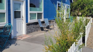 Une maison bleue avec un mur en pierre et une clôture blanche en bois. Il y a deux chaises à l’extérieur et une porte avec une fenêtre.