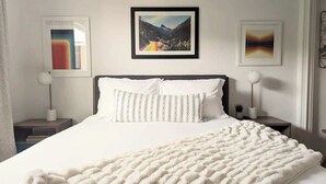 Een net opgemaakt bed met een wit deken en kussens, kunstwerken aan de muur en een foto van een berglandschap boven het bed.