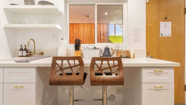 Een keuken met witte kastjes, een dubbele spoelbak, twee houten stoelen en een raam met uitzicht op groen.