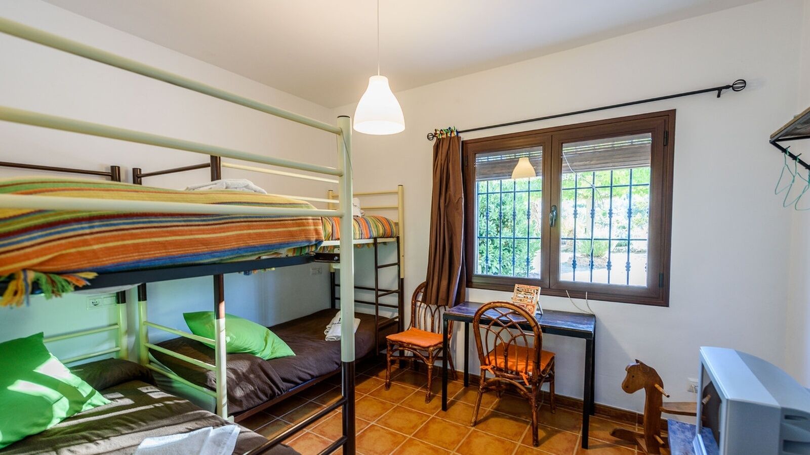 Ein Schlafsaalzimmer mit Etagenbetten, einem Schreibtisch, zwei Stühlen, einem Fenster mit Aussicht und einer Deckenleuchte.