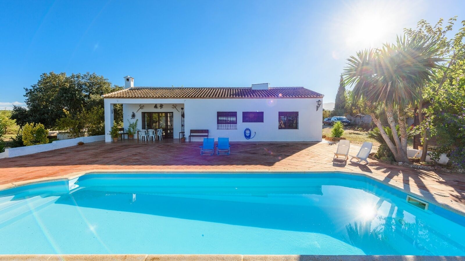 Un espace aménagé au bord d’une piscine à l’eau d’un bleu limpide, un bâtiment blanc avec un toit recouvert de tuiles et une terrasse dotée de chaises et d’une table.