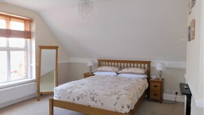 A bedroom with a wooden bed, a nightstand, a lamp, a radiator, and a window with blinds. - Corner House -  a house that sleeps 8 guests  in 4 bedrooms (Sheringham)