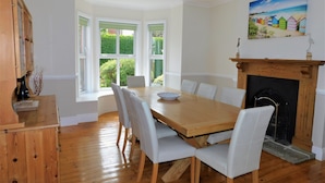A dining room with a wooden table, white chairs, a fireplace, and a painting of a beach scene. - Corner House -  a house that sleeps 8 guests  in 4 bedrooms (Sheringham)