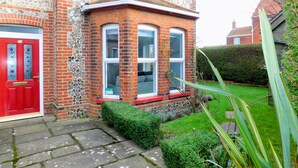 A red door with a number plate, a stone wall, and a brick building with a red window frame and a small porch. - Corner House -  a house that sleeps 8 guests  in 4 bedrooms (Sheringham)