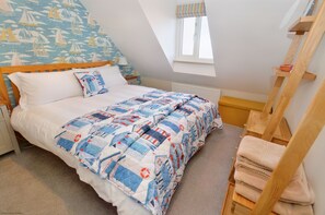 A bedroom with a bed, a wooden headboard, a window with a striped curtain, a wooden ladder, and a radiator. - Gull View - a second floor apartment that sleeps 4 guests in 2 bedrooms (Sheringham)