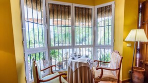 Une pièce d’angle avec une table et deux chaises, de grandes fenêtres avec des barreaux en métal et une lampe.