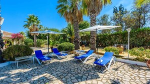 Un espace de détente extérieur baigné de lumière, avec un sol en pierre, des fauteuils bleus et des parasols blancs.
