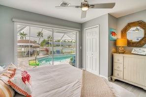 A bedroom with a large bed, a ceiling fan, a view of a swimming pool, and a white dresser with a mirror.