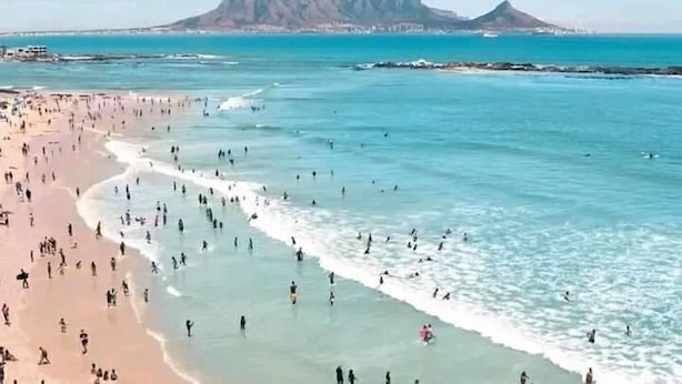 Une plage où l’on voit des gens se baigner et se promener le long du rivage, un ciel dégagé et des montagnes en arrière-plan.