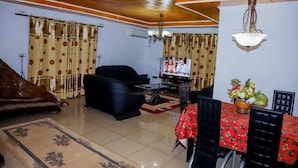 Un salon comprenant un canapé, un fauteuil noir, une table à manger avec des chaises, un téléviseur et un ventilateur de plafond.
