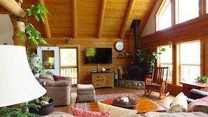 Ein gemütliches Blockhaus-Interieur mit Wohnzimmer, Sofa, Fernseher, Kamin und großen Fenstern.