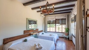Ein Zimmer mit zwei Betten, einer hölzernen Bank, einem Tisch mit einer Lampe und einem Fenster mit Vorhängen.