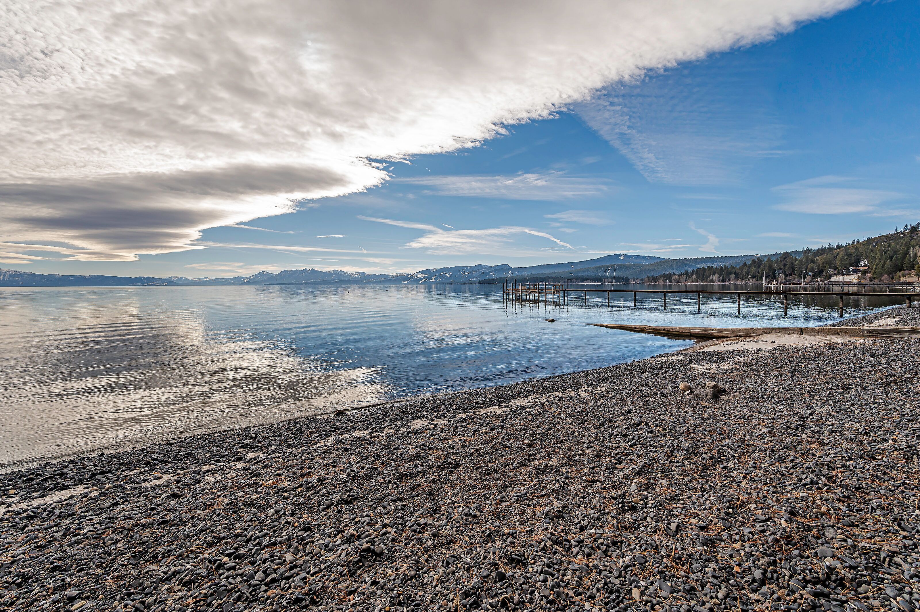 A pebbled beach with a pier extending into calm waters, surrounded by mountains and clear skies.
