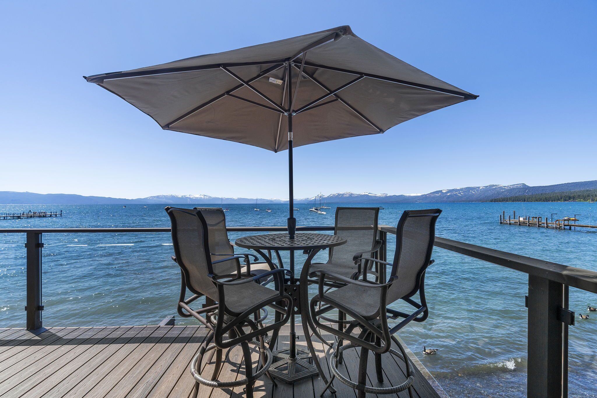 Una terraza con una mesa y sillas bajo una sombrilla, con vistas a un lago con barcos y un muelle.