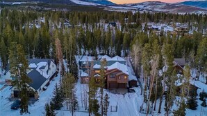 Lumipeitteinen asuinalue, jossa talot ovat tiheän metsän ympäröimiä ja taustalla kohoavat vuoret.