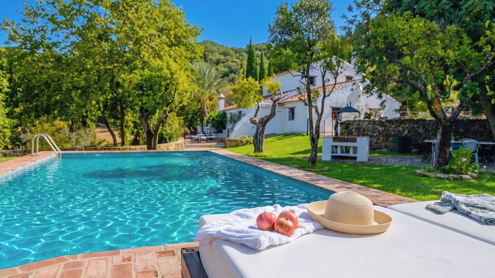 A swimming pool with a white lounge area, a straw hat, and pomegranates on a towel.
