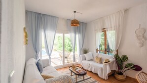 A spacious living room with a sofa, a small table, and a coffee table. There is a large window with curtains, a hanging light fixture, and a dreamcatcher on the wall.