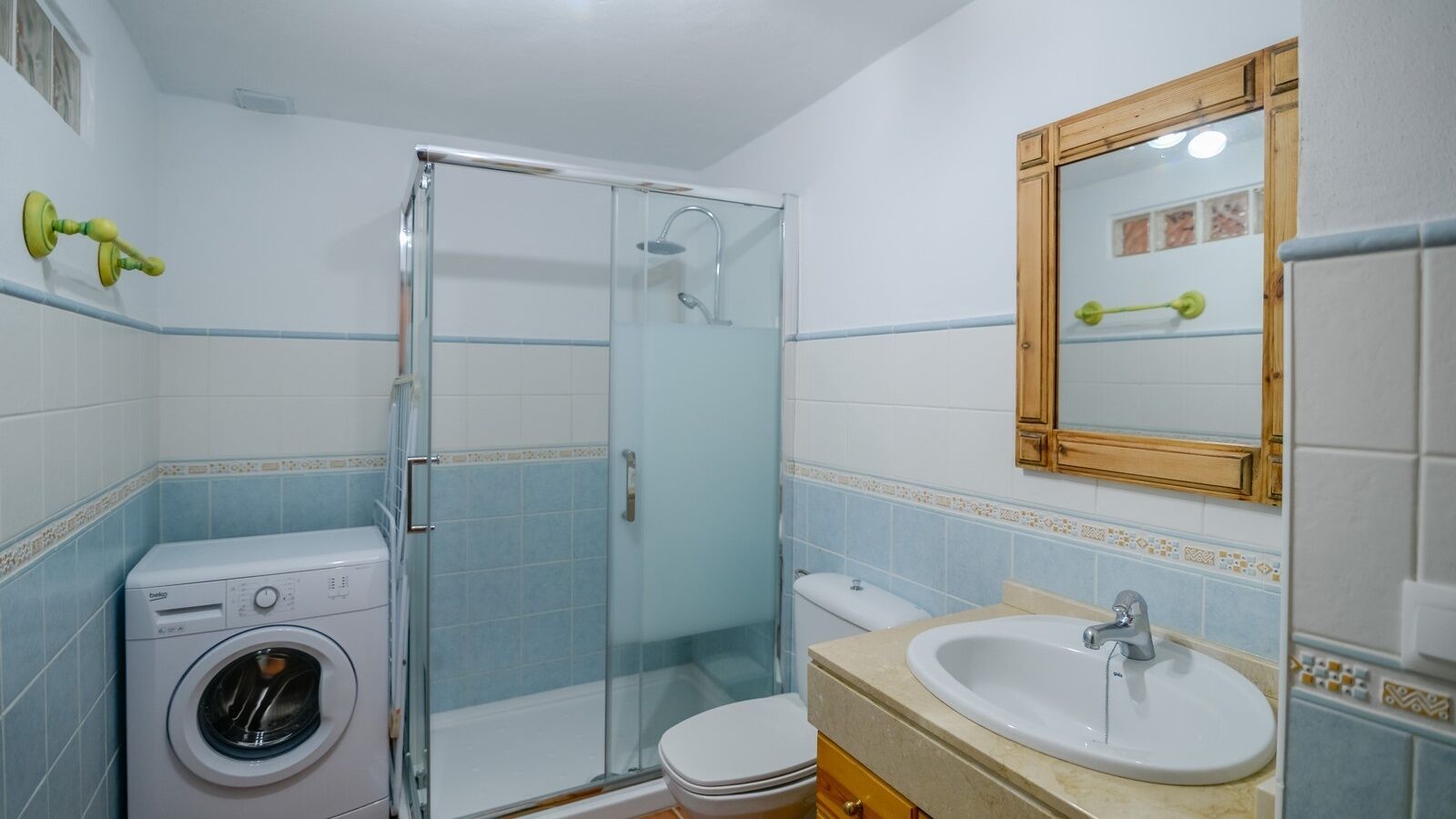 A bathroom with a glass-enclosed shower, a washing machine, a toilet, and a sink with a mirror above it.