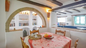A dining area with a round table, chairs, a fruit bowl, and a wall-mounted shelf with books and decorative items.