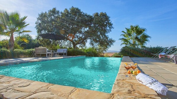 A pool with lounge chairs and a table, surrounded by trees and a clear sky.