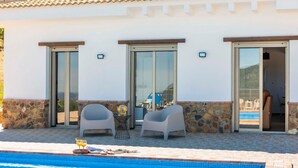 Un espace aménagé au bord de la piscine, avec deux chaises et une table, une piscine, et un bâtiment dont la façade est agrémentée de pierres et qui est doté de portes-fenêtres coulissantes.