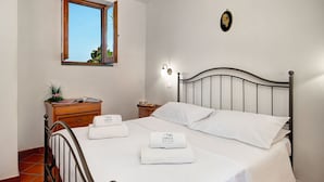 Un lit bien fait, avec du linge de lit blanc et deux serviettes pliées, une commode en bois avec un miroir, un luminaire fixé au mur et une fenêtre donnant sur un paysage verdoyant.