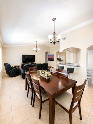 A spacious living room with a dining area, a black leather sofa, a flat-screen TV, and a chandelier.