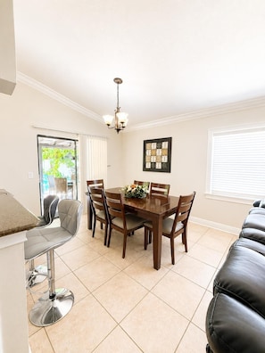 A dining area with a wooden table, six chairs, a chandelier, and a view of greenery outside.