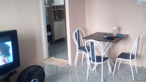 Un petit coin repas avec une table et quatre chaises, un téléviseur et une cuisine visible à travers une porte ouverte.