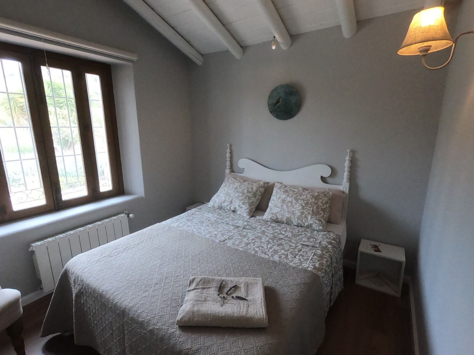 A bedroom with a bed, a window, a radiator, a wall clock, and a bedside table.