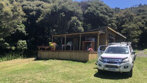 Une cabane avec une terrasse, un camion garé devant, et une colline boisée en arrière-plan.