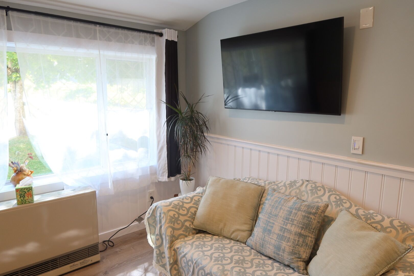 Een woonkamer met een bank, een kamerplant, een radiator en een aan de muur bevestigde televisie.