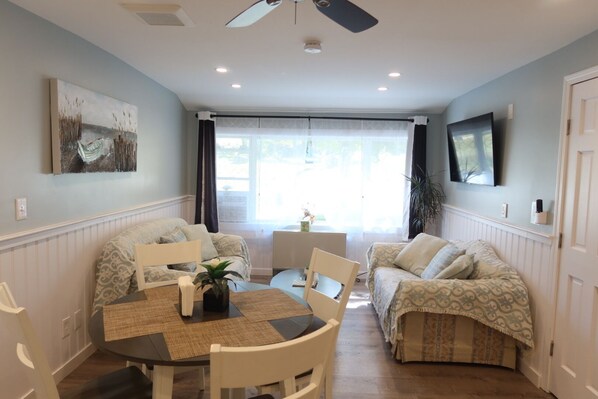 A room with a dining table, two sofas, a ceiling fan, a TV, and a window with curtains.