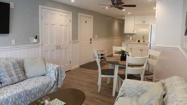A living room with a sofa, a coffee table, and a dining area with chairs and a table. There is a kitchen area with cabinets and a ceiling fan.